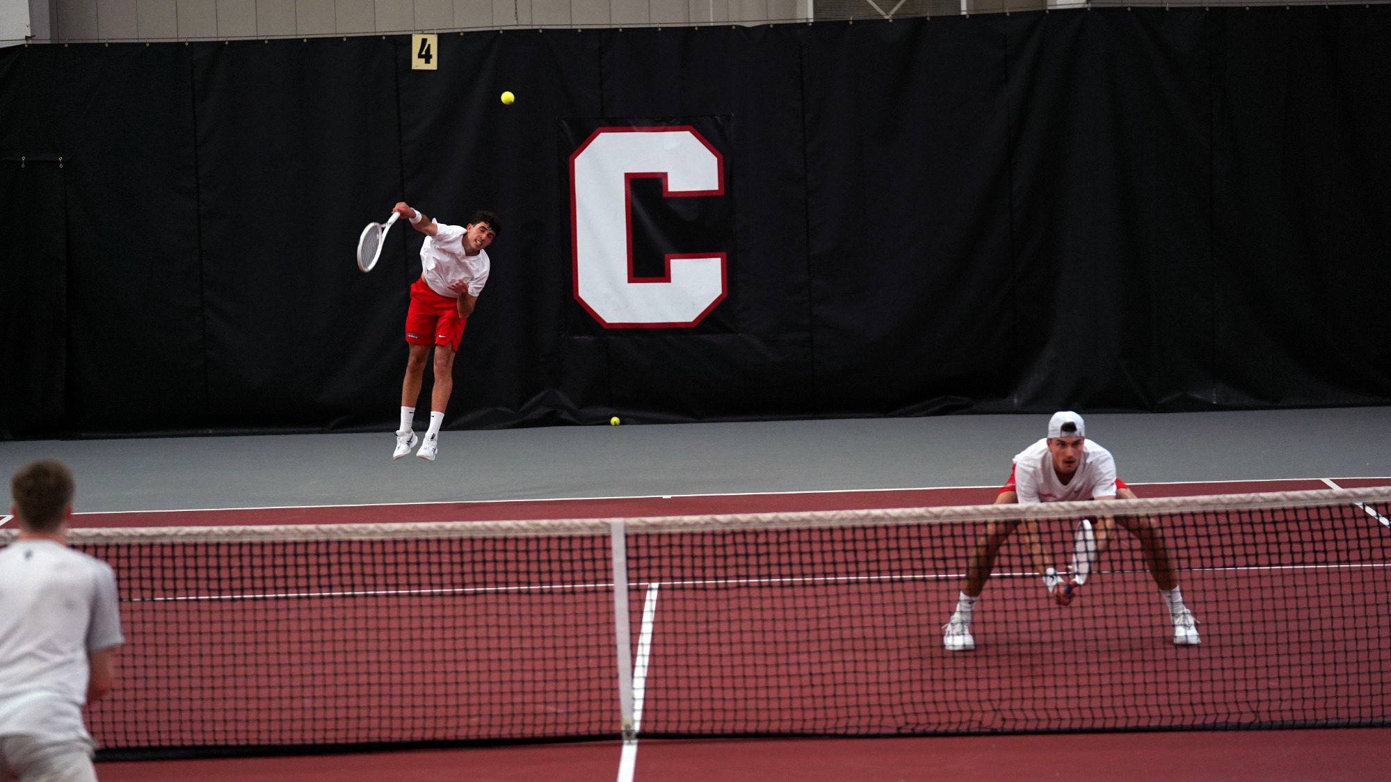 The Cornell men's tennis players compete in doubles with one playing serving and another waiting at the net.