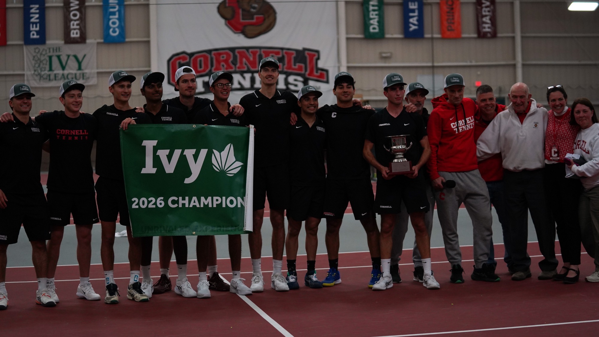 The Cornell men's tennis team celebrates on the court following its 4-3 win over Columbia to win a share of the Ivy League title while holding the team trophy and a white banner that reads Ivy 2026 Champions.