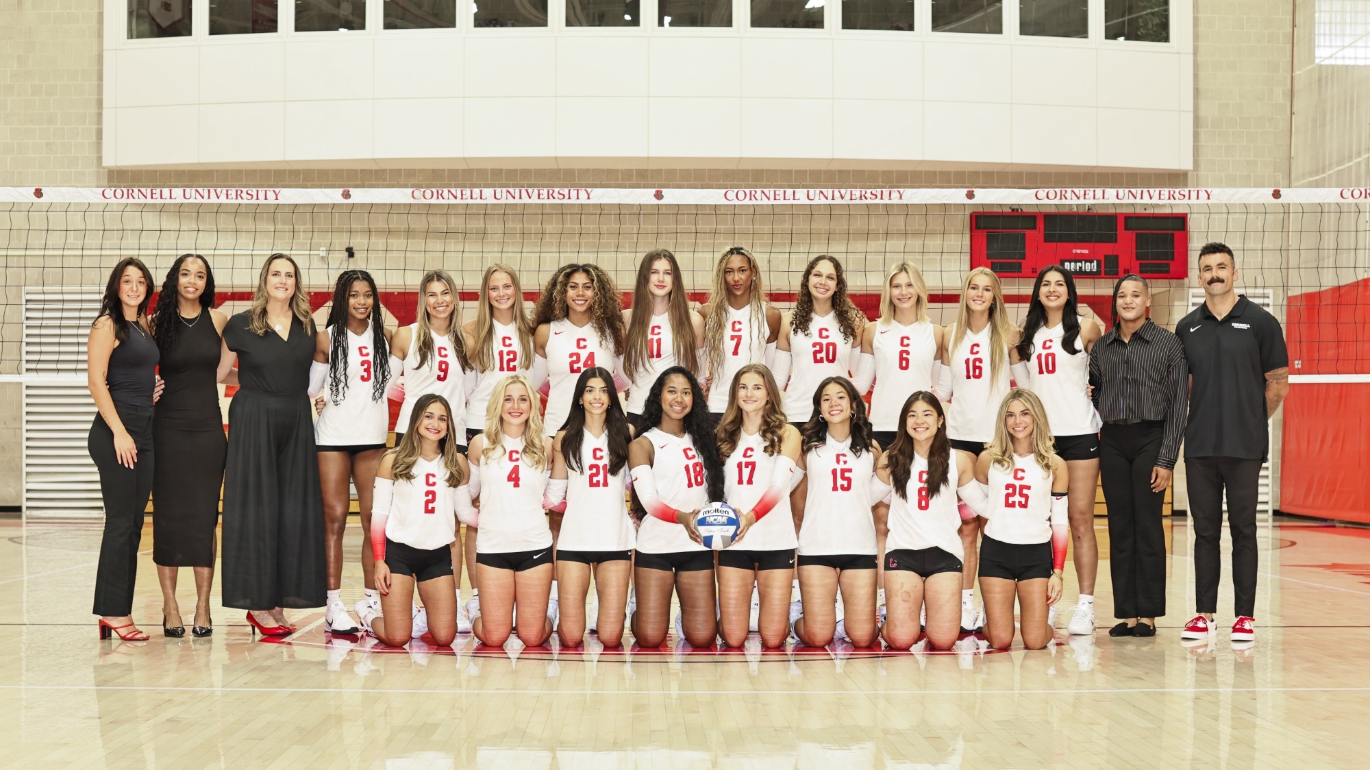 Cornell women’s volleyball team poses for a group photo in uniform on the court, with coaches and staff standing alongside the players.