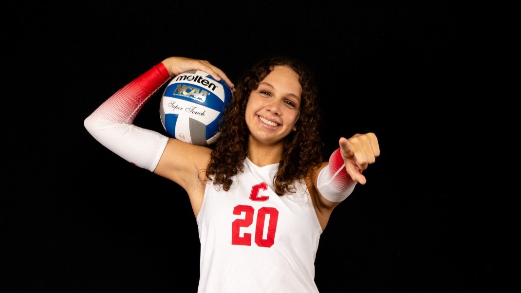 Mackenzie Parsons, no. 20 on Cornell Women's Volleyball posing for Media Day Picture, smiling, volleyball held on her shoulder and other hand pointing at the camera. Wearing white jersey with red no. 20 on the front.