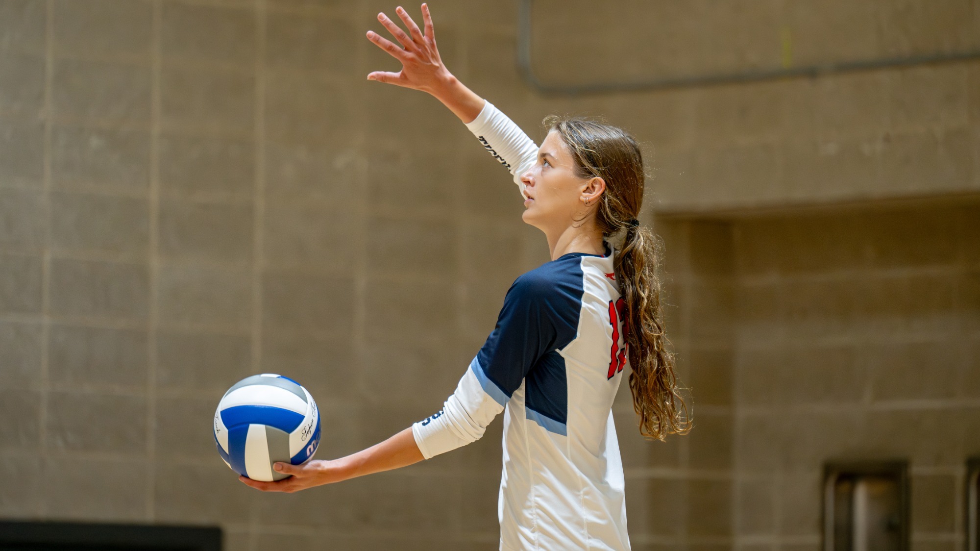 FIT women’s volleyball player Anya Lollos raises one arm while holding a volleyball as she prepares to serve during a match.