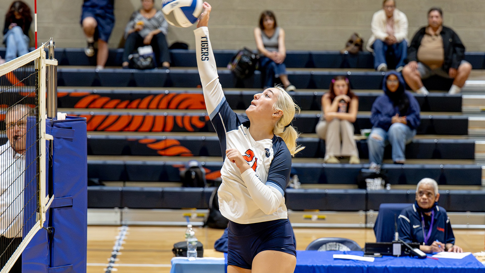 FIT women's volleyball player Brigitte Burns reaches above the net to play the ball during a match, with spectators seated in the gymnasium bleachers behind her.