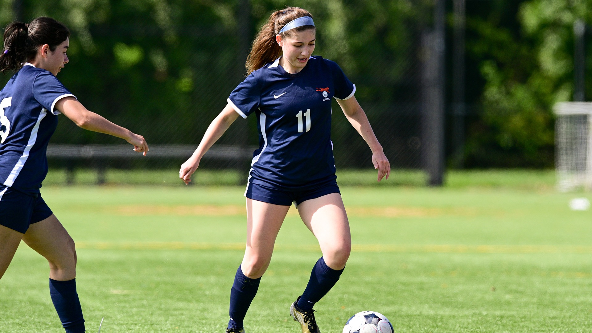 FIT women's soccer player Julia Lothian (#11) controls a soccer ball on the field while wearing a navy blue Tigers uniform and light blue headband. A teammate is positioned beside her during the action.