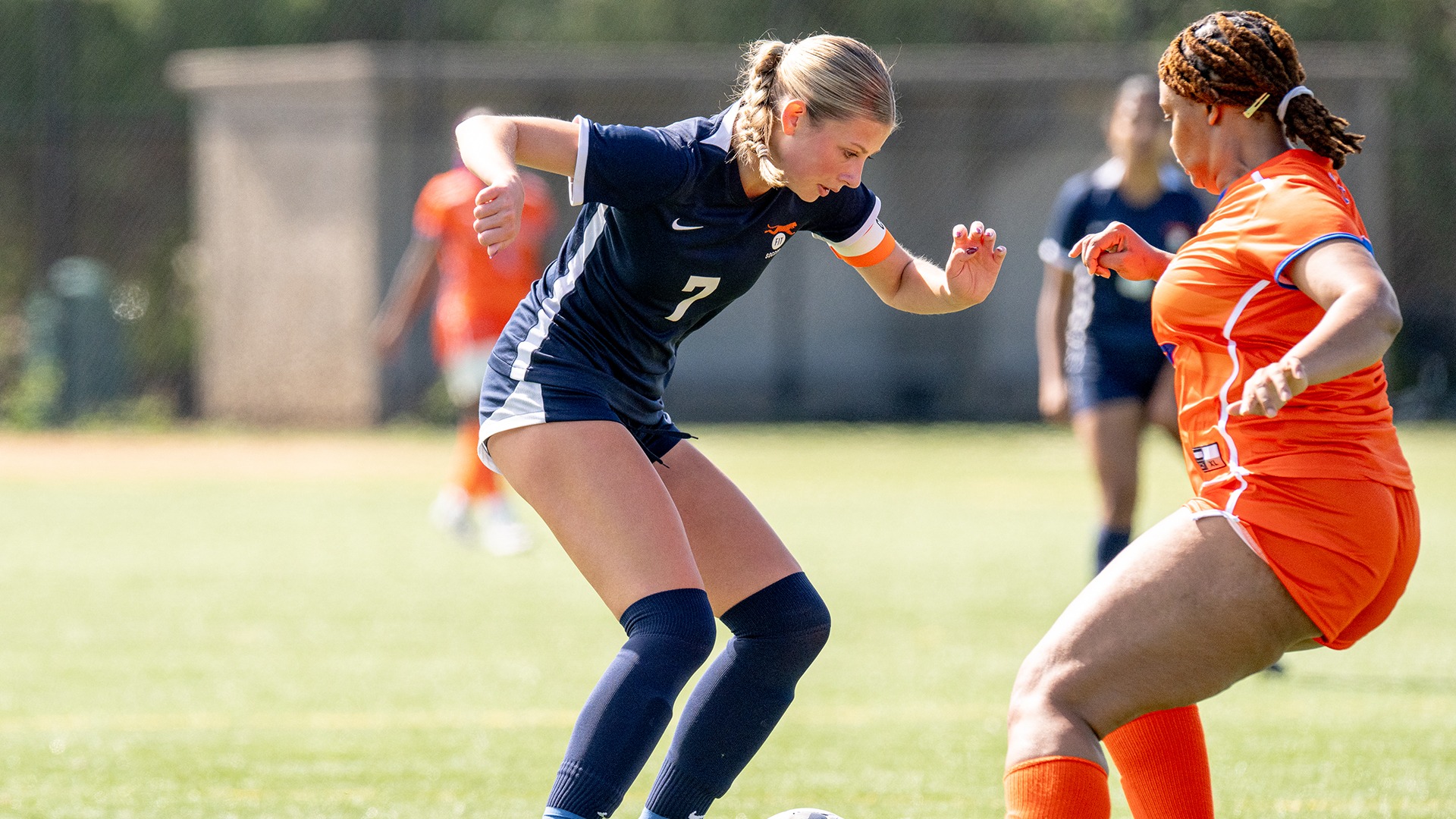 FIT women's soccer defender Kendall Fernaays (#7), wearing a navy blue Tigers uniform and orange captain's armband, battles for possession against an opposing player in an orange uniform during a match.