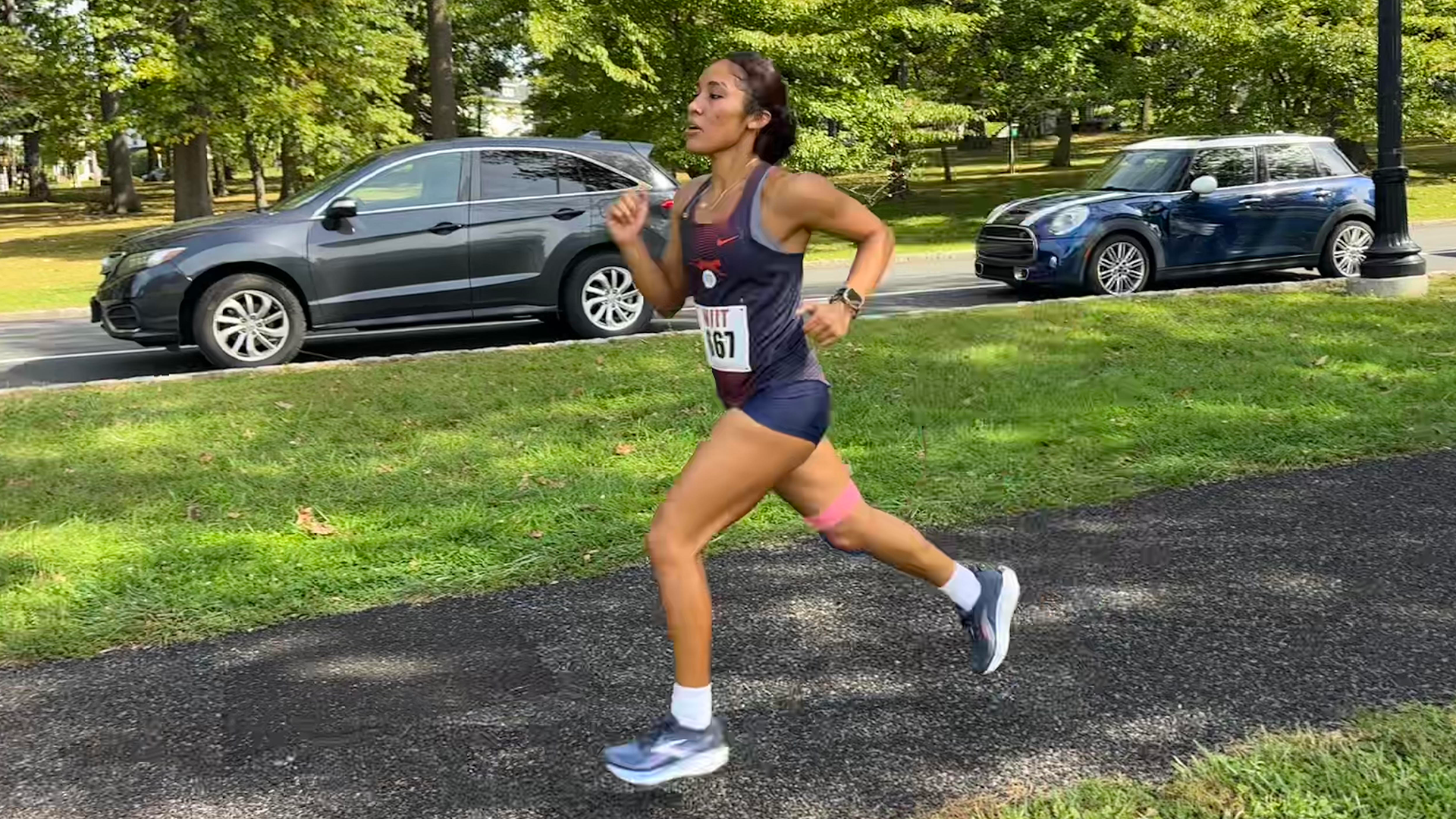 FIT women's cross country runner Mayelie Mena competes in an outdoor race, wearing a navy FIT uniform with bib number 167 as she runs along a paved path bordered by grass and trees.