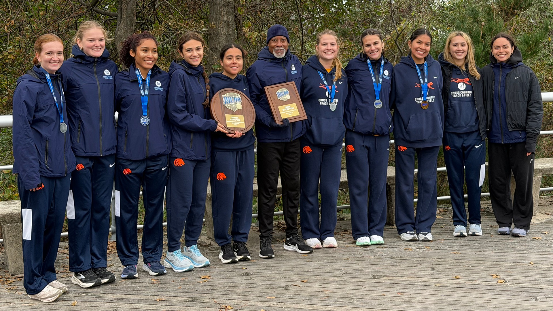 Members of the Fashion Institute of Technology (FIT) women’s cross country team pose with their coach following their third consecutive NJCAA Region 15 Championship at the St. Joseph’s Invitational. The Tigers wear navy blue FIT athletic apparel and display championship plaques and medals while standing together outdoors.