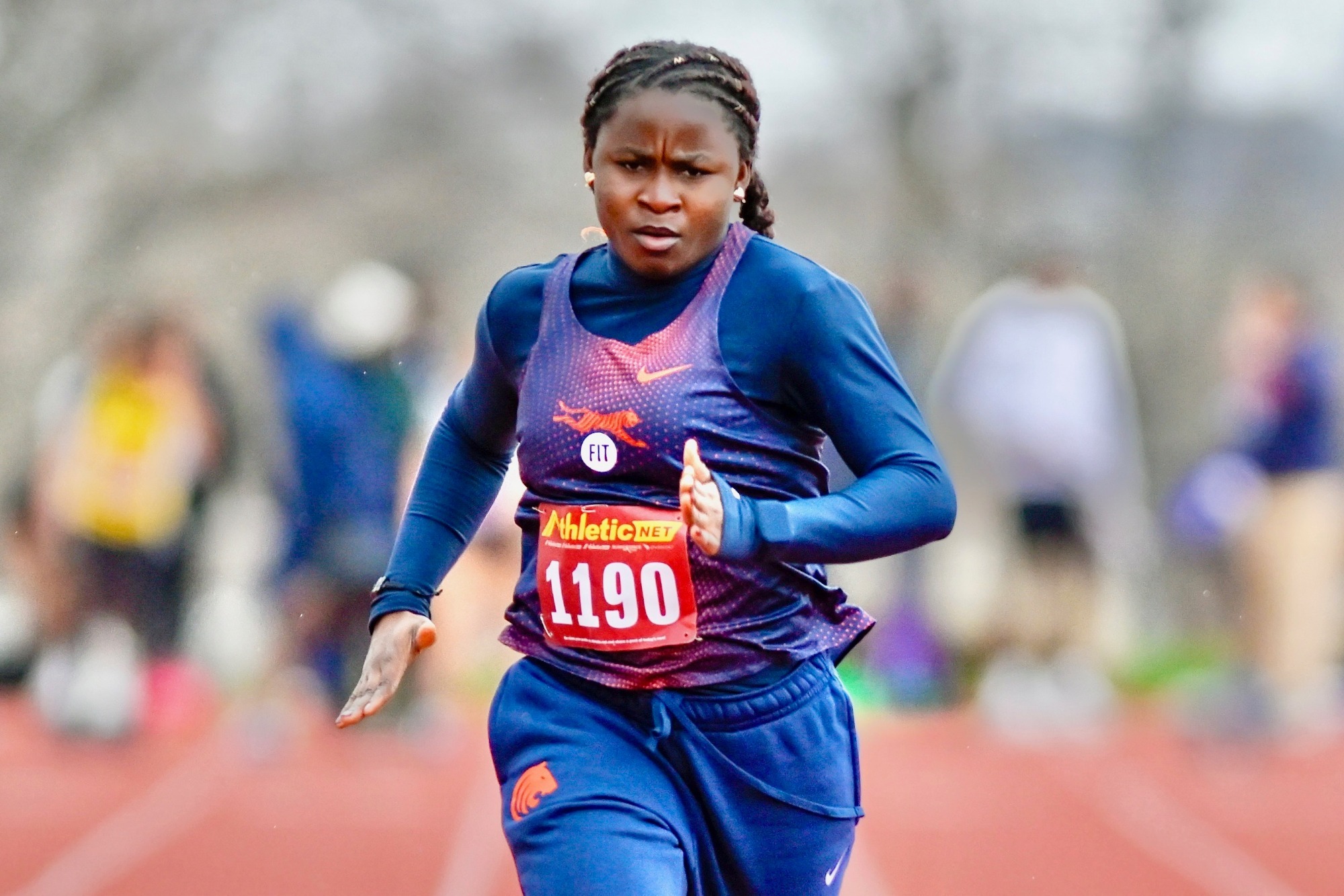 A FIT track athlete wearing a navy uniform with orange accents and bib number 1190 sprints down the track during an outdoor meet, focused mid-stride with blurred competitors and spectators in the background.