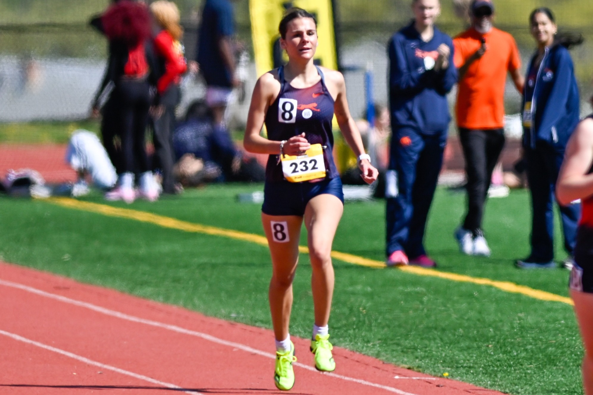 Female track athlete in a navy uniform with bib number 232 running on an outdoor track during a race, with spectators and other competitors visible in the background.