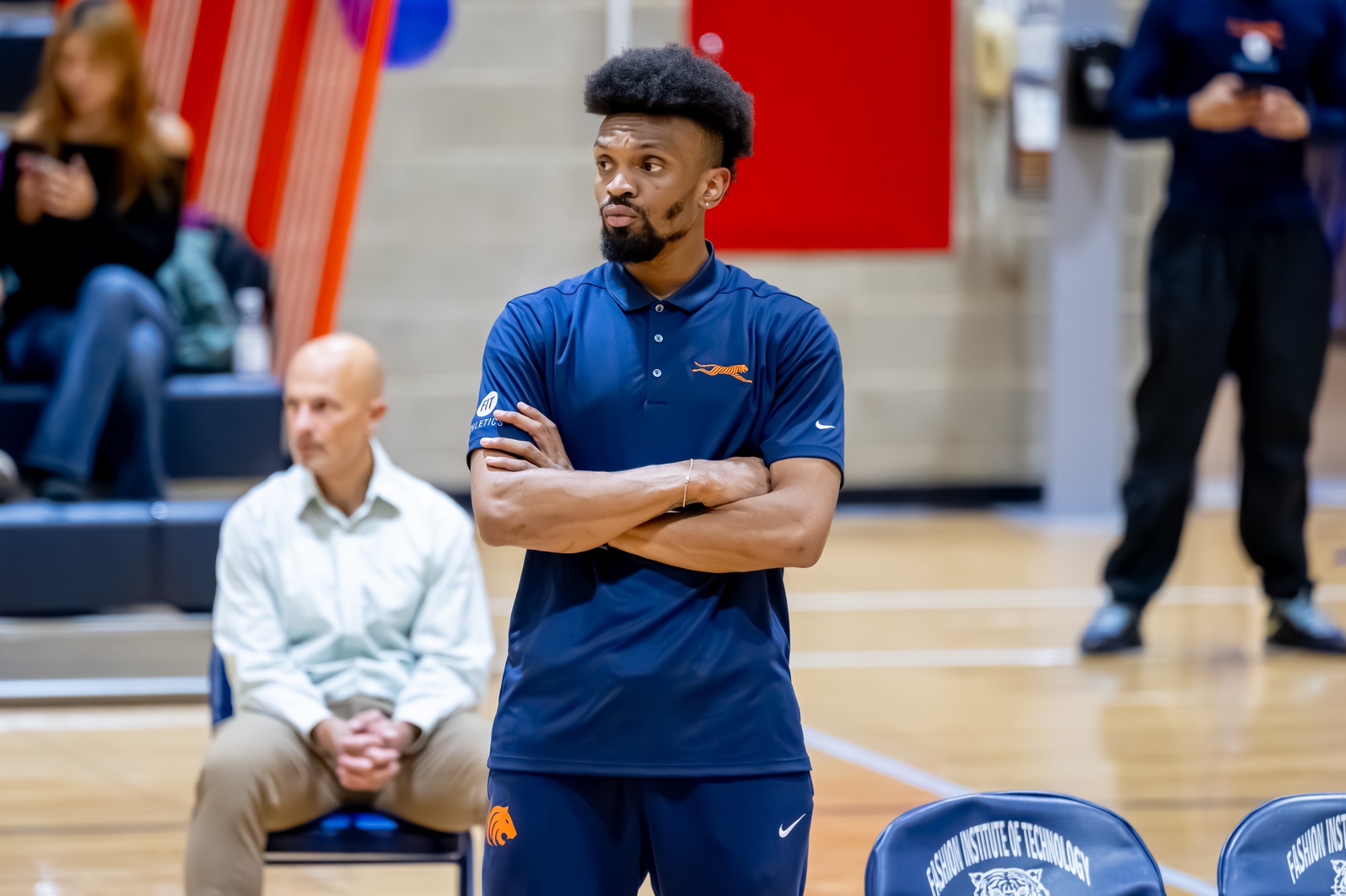Centryll Scott stands courtside with his arms crossed during a volleyball match at the Fashion Institute of Technology. Dressed in FIT volleyball apparel, Scott watches the action from the sideline while spectators and team personnel are visible in the background. The image was taken inside FIT's gymnasium and highlights Scott in his coaching role with the Tigers volleyball program.