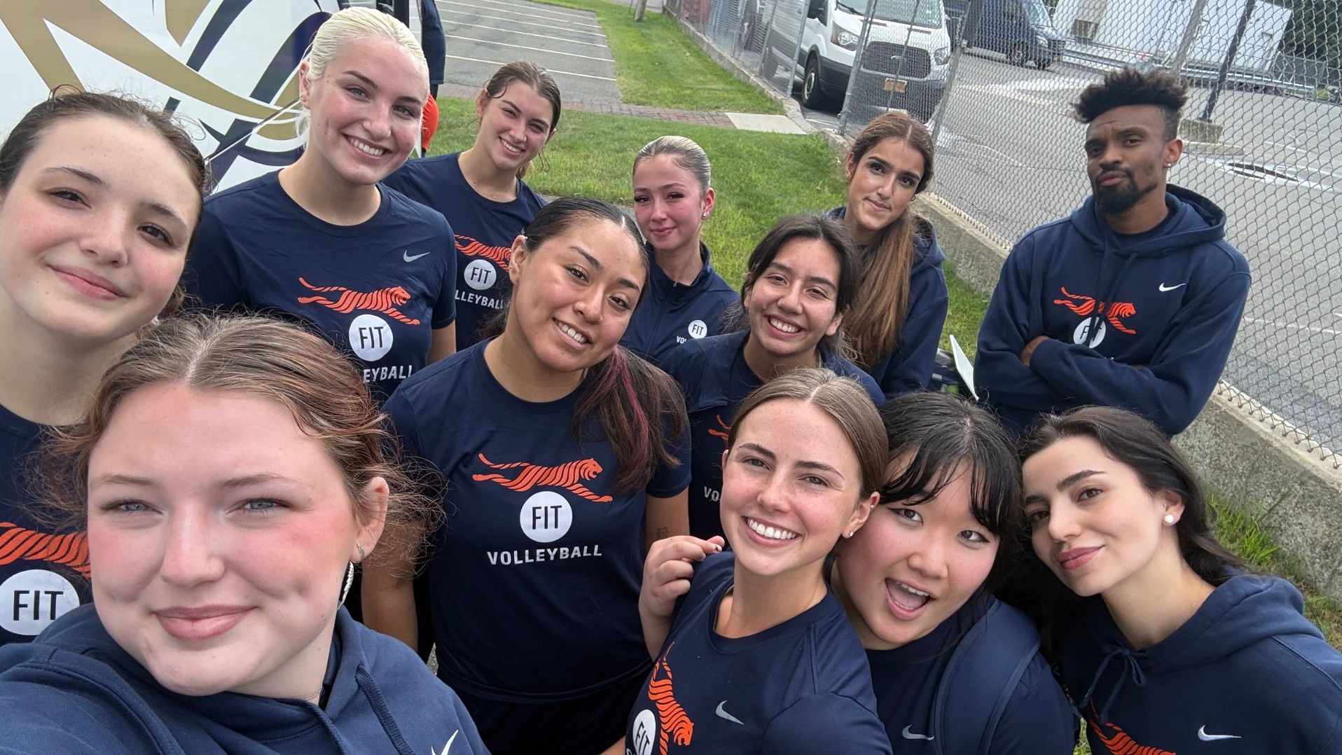 Members of the FIT women’s volleyball team and their coach smile for a group selfie outdoors beside a bus. The team wears navy shirts and hoodies featuring the orange FIT Tigers logo.