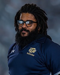 Gallaudet men & women's track and field 2023 headshot Ronald Mattingly. The coach is wearing a navy blue polo shirt with a GU Bison Logo on it. The coach is photographed with a gray background in the back.