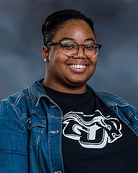 Gallaudet winter cheerleading 2022-2023 headshot Shea Jefferson. The coach is wearing a denim jacket and black shirt with a GU Bison Logo on it. The coach is photographed with a gray background in the back.