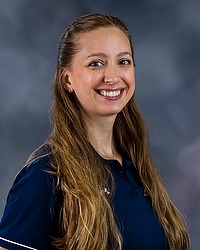 2023 WVB Headshot - Ann Behm - Women's volleyball assistant coach looks towards the camera for a headshot. The coach is wearing a blue collared Bison shirt. There is a gray backdrop behind the coach.