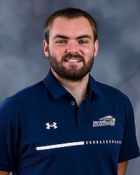2023 WVB Headshot - Ben Campman - Women's volleyball graduate assistant looks towards the camera for a headshot. The assistant is wearing a blue collared Bison shirt. There is a gray backdrop behind the assistant.