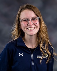 Gallaudet Track & Field 2024 headshot Sierra Pratt. The coach is wearing a navy blue pullover with the word GALLAUDET TRACK & FIELD on chest. The coach is photographed with a gray background in the back