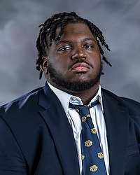 Bobby Battle 2024 Gallaudet University football headshot. Football player looks towards the camera for a headshot. The player is wearing a white collared shirt with a blue bison tie under a navy blue blazer. There is a gray backdrop behind the player.