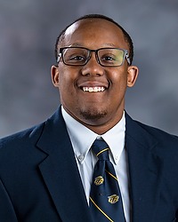 Joshua Huff 2024 Gallaudet University football headshot. Football player looks towards the camera for a headshot. The player is wearing a white collared shirt with a blue bison tie under a navy blue blazer. There is a gray backdrop behind the player.