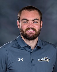 2024 WVB Headshot - Ben Campman - Women's volleyball assistant coach looks towards the camera for a headshot. The coach is wearing a blue collared Bison shirt. There is a gray backdrop behind the coach.