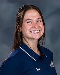 2024 WVB Headshot -Leila Sicoli - Women's volleyball assistant coach looks towards the camera for a headshot. The coach is wearing a blue collared Bison shirt. There is a gray backdrop behind the coach.