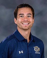 2024 MSOC Headshot - James Rummo - Men's soccer assistant coach looks towards the camera for a headshot. The assistant coach is wearing a blue Bison collared shirt. There is a gray backdrop behind the coach.