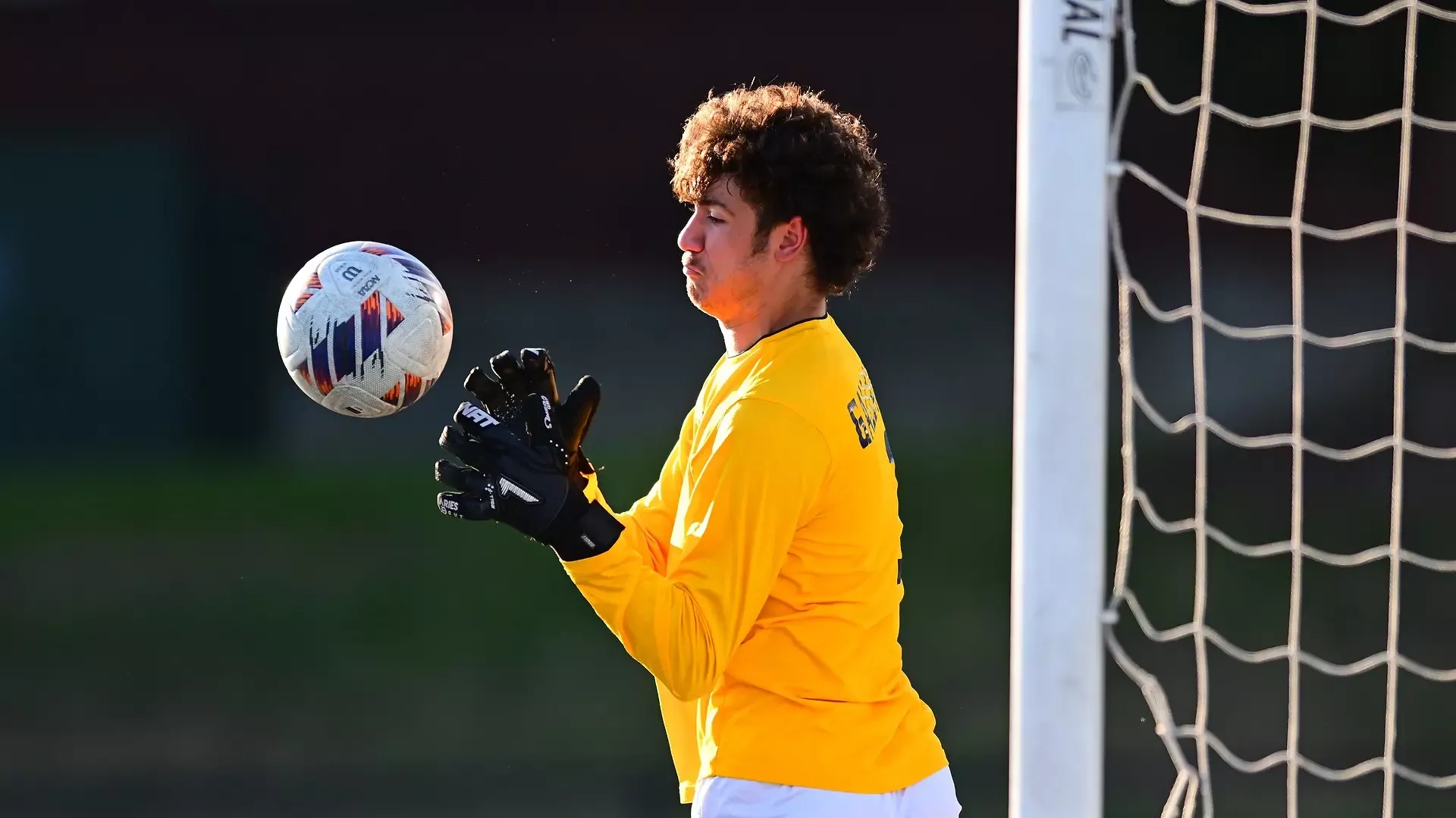 Gallaudet men's soccer goalie John McCabe makes a save during a late afternoon game as the sun sets in the distance. McCabe is wearing a yellow/gold goalie jersey and black goalie gloves.