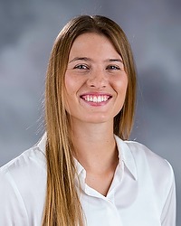 Cam Lexow - 2025-26 Gallaudet women’s basketball headshot. The student coach wears a white blouse and looks towards the camera. A gray backdrop is in the background.