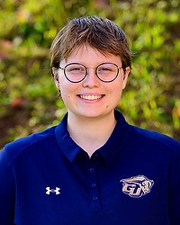 Mara Land- 2025-26 Gallaudet men & women’s swim headshot. The Bison assistant coach wears a navy blue polo shirt and looks towards the camera. Trees are in the background.