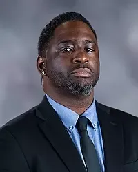 Gallaudet University men's basketball assistant coach Kazeem Babatunde headshot photo. He is wearing a black colored blazer, a blue button down shirt and blue tie against a grey background.
