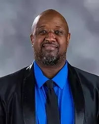 Gallaudet University men's basketball life coach Reginald Bess headshot photo. He is wearing a black colored blazer, a blue button down shirt and black tie against a grey background.