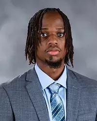 Gallaudet University men's basketball assistant coach Blessed Mbogo headshot photo. He is wearing a gray colored blazer, a white button down shirt and blue tie against a grey background.