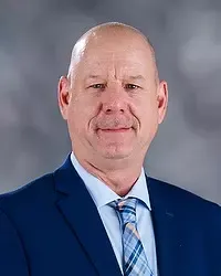 Gallaudet University men's basketball assistant coach Keith Westhoelter headshot photo. He is wearing a blue colored blazer, a white button down shirt and blue tie against a grey background.