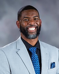 Gallaudet University men's basketball head coach Sekoe White headshot photo. He is wearing a grey colored blazer, a black button down shirt and blue tie against a grey background. 