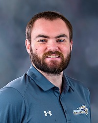 2025 MVB Headshot - Ben Campman- Men's volleyball coach looks towards the camera for a headshot. The coach is wearing a grey Bison polo shirt. There is a gray backdrop behind the player.