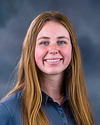 2025 MVB Headshot - Ella Gamble- Men's volleyball coach looks towards the camera for a headshot. The coach is wearing a grey Bison polo shirt. There is a gray backdrop behind the player.