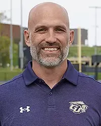 Gallaudet football head coach Stefan LeFors headshot taken on Hotchkiss Field on an overcast day. LeFors is wearing a navy blue polo and is smiling at the camera.