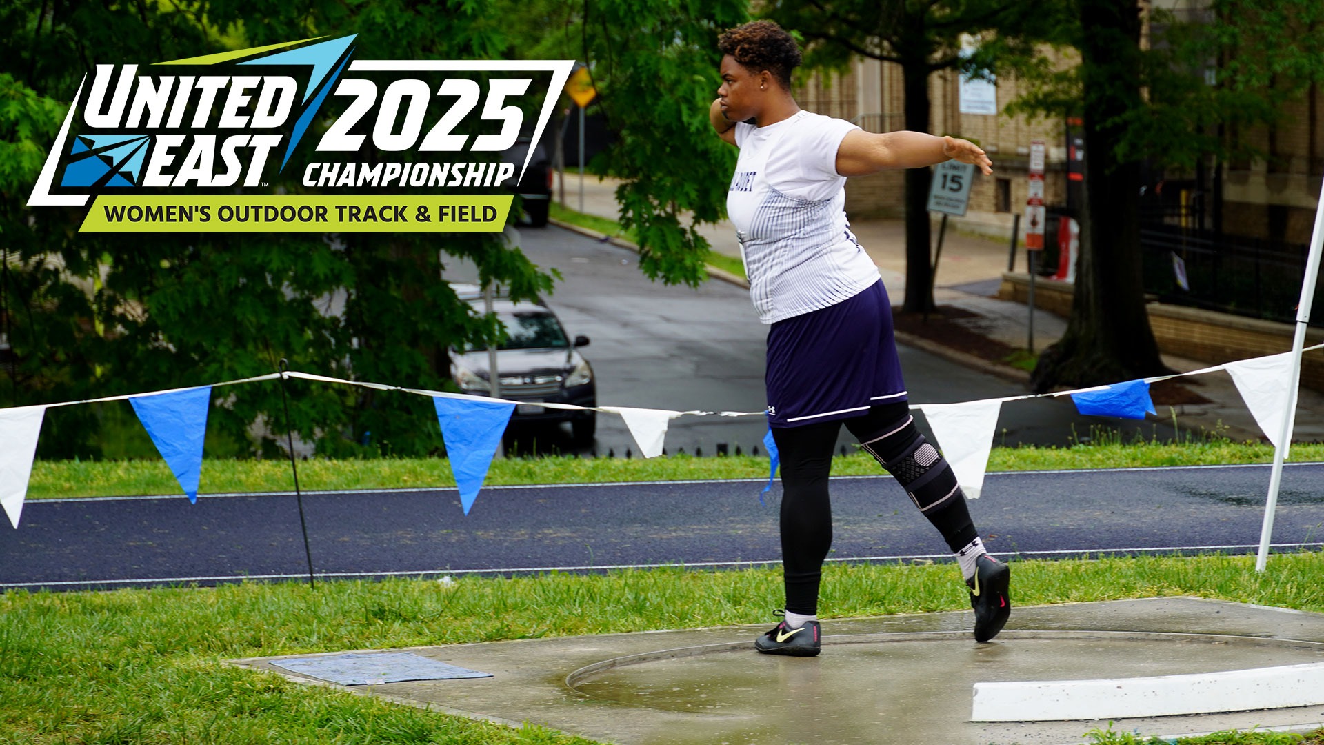 Gallaudet women's track and field thrower Midajah Davis prepares to throw the silver shot put during an overcast and wet day in the nation's capital. A 2025 United East Women's Outdoor Track and Field logo is in the upper left corner.