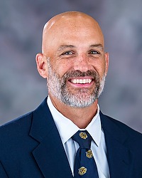Stefan LeFors 2025 Gallaudet University football headshot. Football head coach looks towards the camera for a headshot. The coach is wearing a white collared shirt with a blue bison tie under a navy blue blazer. There is a gray backdrop behind the coach.