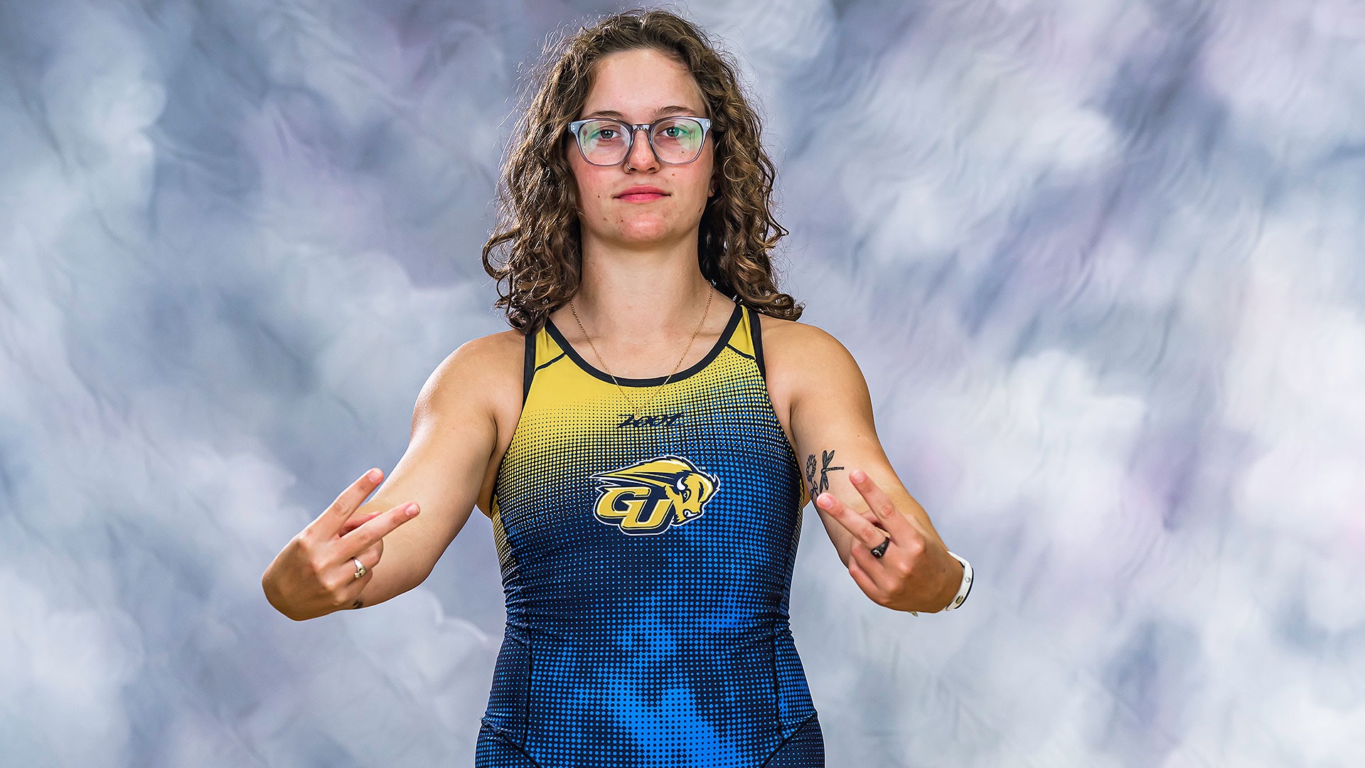 Gallaudet triathlete Ashley Corcoran stands in front of a gray background in a triathlon suit and makes the peace sign with both hands.