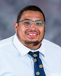 Aubrey Moorman 2025 Gallaudet University football headshot. Football coach looks towards the camera for a headshot. The coach is wearing a white collared shirt with a blue bison tie under a navy blue blazer. There is a gray backdrop behind the coach.