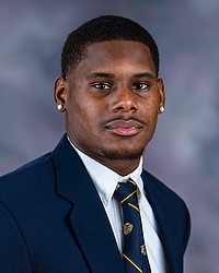 Brandon Washington 2025 Gallaudet University football headshot. Football coach looks towards the camera for a headshot. The coach is wearing a white collared shirt with a blue bison tie under a navy blue blazer. There is a gray backdrop behind the coach.
