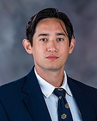 Jonah Wooley 2025 Gallaudet University football headshot. Football coach looks towards the camera for a headshot. The coach is wearing a white collared shirt with a blue bison tie under a navy blue blazer. There is a gray backdrop behind the coach.