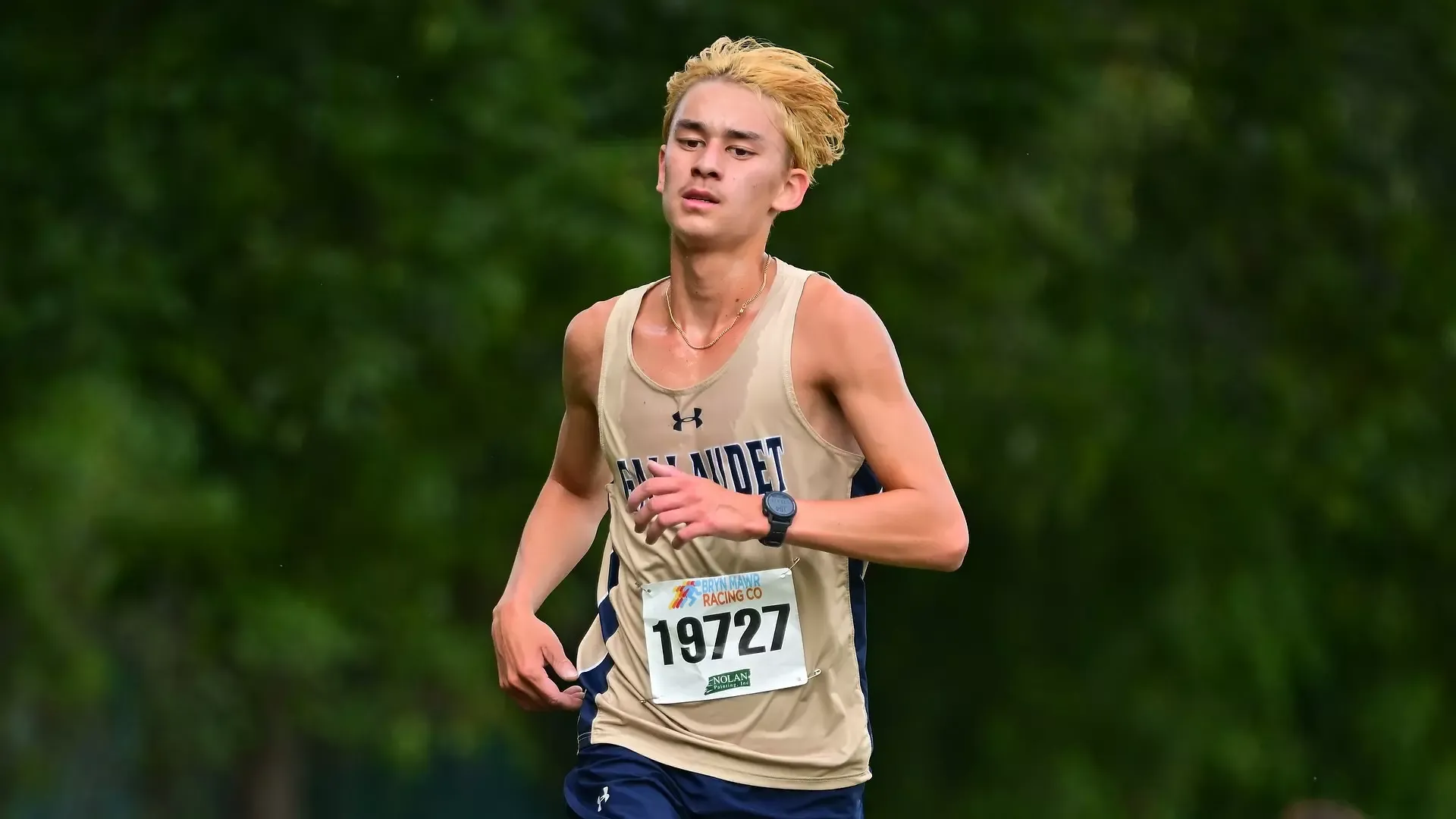 Gallaudet men's cross country runner Sevan Ikeda runs on a sunny day in a cross country meet. This photo is a solo photo of him with trees in the background.