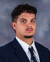 Gabe Segovia 2025 Gallaudet University football headshot. Football player looks towards the camera for a headshot. The player is wearing a white collared shirt with a blue bison tie under a navy blue blazer. There is a gray backdrop behind the player.
