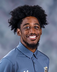 2025 MSOC Headshot - Kelly Grant - Men's soccer coach looks towards the camera for a headshot. The coach is wearing a grey polo shirt. There is a gray backdrop behind the player.
