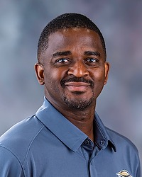 2025 MSOC Headshot - Christopher Jappah - Men's soccer coach looks towards the camera for a headshot. The coach is wearing a grey polo shirt. There is a gray backdrop behind the player.