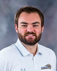 2025 WVB Headshot - Ben Campman Women's volleyball coach looks towards the camera for a headshot. The coach is wearing a white polo shirt. There is a gray backdrop behind the player.