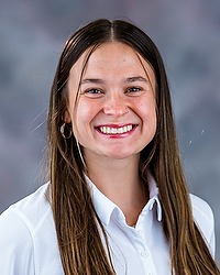 2025 WVB Headshot - Leila Sicoli Women's volleyball coach looks towards the camera for a headshot. The coach is wearing a white polo shirt. There is a gray backdrop behind the player.