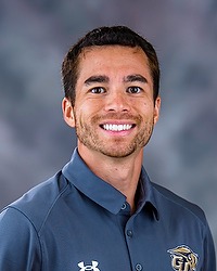 2025 MSOC Headshot - James Rummo - Men's soccer coach looks towards the camera for a headshot. The coach is wearing a grey polo shirt. There is a gray backdrop behind the coach.