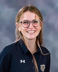 2025 XC Headshot - Sierra Pratt - men's & women's cross country coach looks towards the camera for a headshot. The coach is wearing a black polo shirt. There is a gray backdrop behind the coach.