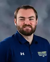 Ben Campman - 2026 Gallaudet men’s volleyball headshot. The Bison interim head coach wears a navy colored polo and looks towards the camera. A gray backdrop is in the background.